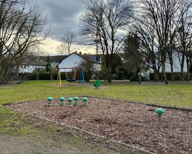 Im Hintergrund stehen weiße Häuser, davor hohe, kahle Bäume, man sieht einen Spielplatz mit Rasen, einer Nestschaukel und einem Sandkasten.